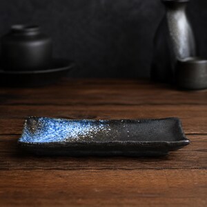 Op de afbeelding: Een rechthoekig keramisch bord met een getextureerd zwart oppervlak en een verloop van gespikkelde blauwe en witte glazuur aan één kant. Het bord heeft opstaande randen en een rustieke, onregelmatige afwerking die het contrast benadrukt.