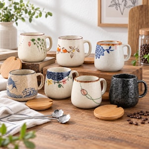 May include: A collection of seven ceramic mugs with speckled finishes, featuring various hand-painted designs including birds, flowers, and waves. Each mug includes a matching wooden lid, and two silver-toned spoons with floral-shaped handles.