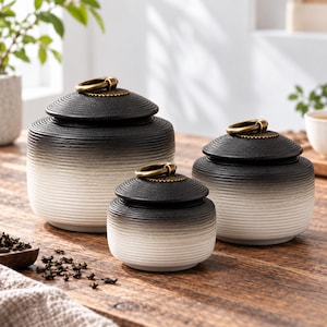 Puede incluir: Tres tarros de almacenamiento de cerámica con tapa, con una textura acanalada horizontal y un acabado degradado de negro a blanco. Cada tapa está rematada con un anillo metálico de color latón que sirve como asa para abrir los recipientes.