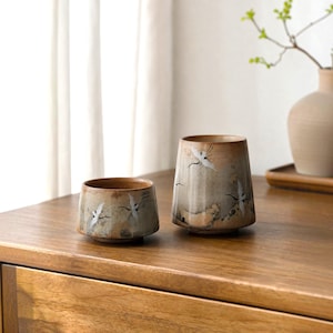 May include: Two ceramic cups with a speckled tan and grey glaze, featuring hand-painted white and black crane motifs. The shorter cup is wide and cylindrical, while the taller cup has a slightly tapered shape with a wider base and narrow rim.