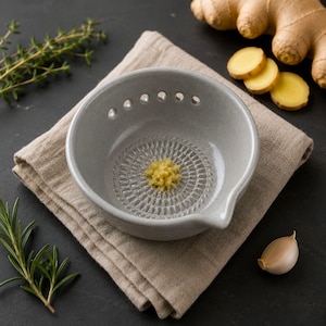 Può includere: Una ciotola grattugia in ceramica grigio chiaro con una superficie di grattugia circolare testurizzata sul fondo e un piccolo beccuccio. La ciotola presenta una fila di piccoli fori lungo il bordo interno e contiene zenzero grattugiato.