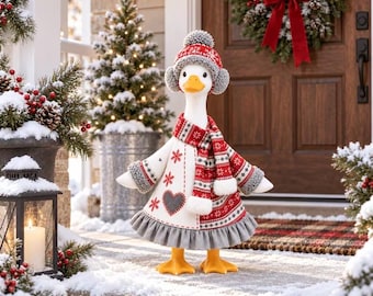 May include: A white goose figurine wearing a red and grey knit hat with a pom-pom, a matching scarf, and a coat featuring snowflake patterns and a grey heart. The goose has an orange beak and orange feet, standing on a snowy porch surface.