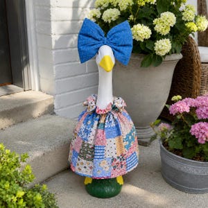 May include: A white goose statue wearing a large blue fabric bow on its head and a ruffled dress featuring a multi-colored patchwork pattern of floral, gingham, and polka dot prints. The goose has a bright yellow beak and stands on a green base.