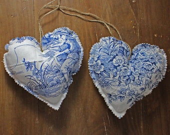 Op de afbeelding: Twee blauwe en witte stoffen hartvormige ornamenten met een bloemenpatroon. De harten hangen aan een touwtje.