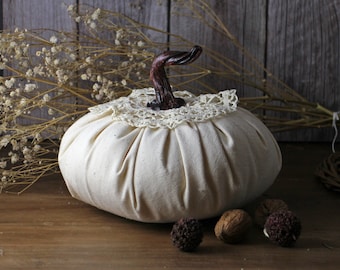May include: A cream-colored fabric pumpkin with a dark brown stem and a cream lace collar. The pumpkin is sitting on a wooden surface with dried flowers and walnuts in the background. A decorative fall item.