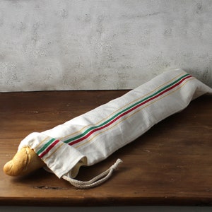 Może przedstawiać: Biały lniany worek na chleb w czerwone, zielone i żółte paski. Worek jest związany białym sznurkiem i zawiera bochenek chleba.