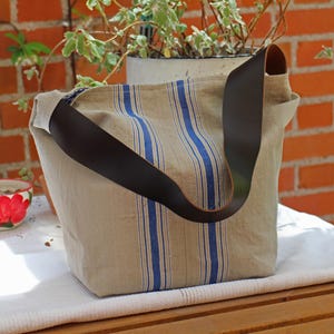 May include: A beige tote bag with vertical blue stripes and a dark brown leather shoulder strap. The bag is made of a textured fabric. The bag is on a white surface. A brick wall and potted plants are in the background.