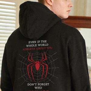 Könnte beinhalten: Ein schwarzer Kapuzenpullover mit einer gestickten roten Spinne in der Mitte eines grauen Spinnennetz-Designs auf der Rückseite. Der Text lautet: EVEN IF THE WHOLE WORLD FORGETS ABOUT YOU DON'T FORGET WHO YOU ARE in weißer und roter Schrift.