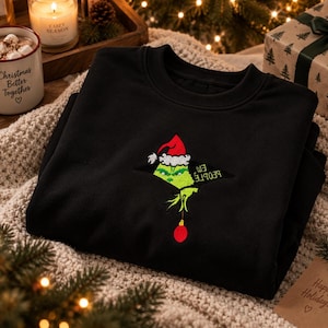 Peut inclure: Un sweat-shirt noir à col rond avec une broderie représentant un personnage vert portant un bonnet de Père Noël rouge, sortant d'une ouverture triangulaire. Le personnage tient une boule de Noël rouge et le texte "EW PEOPLE" est brodé.