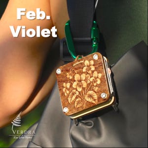 May include: A square wooden accessory featuring a laser-engraved violet flower design, secured with four silver-toned metal screws. The item is attached to a black strap via a bright green carabiner. The text "Feb. Violet" is visible at the top.