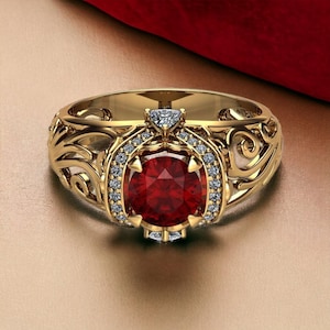 May include: A gold ring featuring a large, round, deep red center stone surrounded by a halo of small, clear, sparkling stones. The band is crafted with intricate, swirling filigree metalwork that extends along the sides of the ring.
