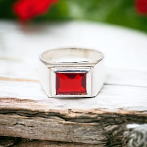 Peut inclure: Une bague en argent ornée d'une pierre précieuse rouge foncé rectangulaire et facettée, montée sur un anneau en métal lisse et poli. La bague présente une large tige solide qui s'affine légèrement vers l'arrière, au design géométrique.