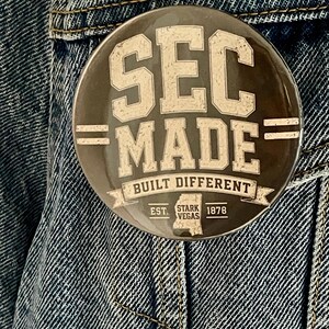 May include: A circular pinback button with a dark background and white distressed text that reads SEC MADE, BUILT DIFFERENT, EST. 1878, and STARK VEGAS, featuring a small map outline of Mississippi at the bottom, attached to a blue denim fabric.