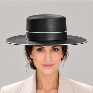 May include: A black flat-top hat with a wide, stiff brim and a smooth, leather-like finish. The hat features a thin, white piping detail that encircles the base of the crown and runs along the outer edge of the brim, creating a clean, structured look.