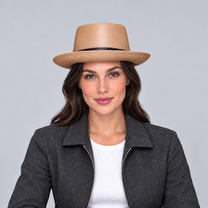 Pode incluir: Um chapéu fedora bege com copa plana e aba larga, apresentando uma faixa fina castanho-escura em torno da base da copa. O chapéu é feito de um material liso e de cor sólida, sendo usado sobre cabelos castanhos escuros.