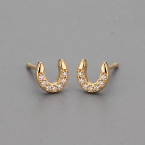 Könnte beinhalten: Ein Paar hufeisenförmige Ohrstecker aus Gold, die jeweils mit einer Reihe kleiner, klarer, rund geschliffener Steine besetzt sind, die entlang der gewölbten Oberfläche verlaufen. Die Ohrringe haben eine polierte metallische Oberfläche.