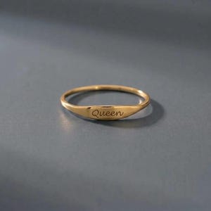 Op de afbeelding: Een dunne goudkleurige ring met een plat, horizontaal gedeelte aan de voorzijde. Het woord Queen is in een cursief lettertype gegraveerd in het midden van de platte balk. De ring is glad, gepolijst en heeft een rond ontwerp.