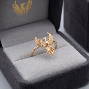 May include: A rose gold ring featuring a detailed phoenix bird with outstretched wings and a flowing tail. The bird is centered on the thin band, showcasing textured feather details across its body and wings, creating a cohesive metallic design.