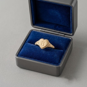 Op de afbeelding: Een gouden ring met een hartvormig oppervlak waarin de letter B in het midden is gegraveerd. De ring heeft een kralenrand en gedetailleerd filigraanwerk langs de band, gepresenteerd in een donkergrijze doos met een diepblauwe fluwelen voering.
