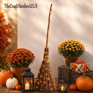 May include: A decorative broomstick made of natural twigs with integrated warm white fairy lights, standing upright. The text TheCraftHarborUSA is visible in the upper left corner. The broom is surrounded by orange and white pumpkins, potted chrysanthemums, and two black metal lanterns with lit candles.