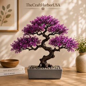 May include: An artificial bonsai tree with vibrant purple leaves and a dark, textured, gnarled trunk. The tree is planted in a rectangular gray pot filled with small light-colored pebbles. The text TheCraftHarborUSA is visible above the tree.