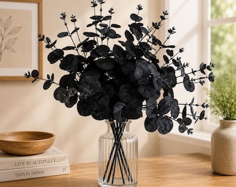 May include: A bouquet of artificial black eucalyptus stems with round leaves arranged in a clear, vertically ribbed glass vase. The stems feature multiple branches with dark foliage, creating a dense, rounded display of dark botanical elements.