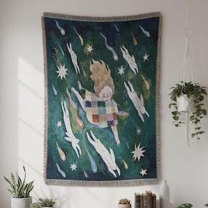 Puede incluir: Un tapiz de pared tejido que presenta una figura central de cabello largo con una túnica de cuadros multicolor, rodeada de conejos blancos y estrellas fugaces de colores sobre un fondo verde oscuro, con un borde de flecos.