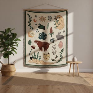 May include: A cream-coloured woven wall tapestry featuring a dark green border and fringe. The design includes various illustrations such as a brown bear, a grey rabbit, a cat face, two human figures, and assorted botanical elements like leaves and flowers.