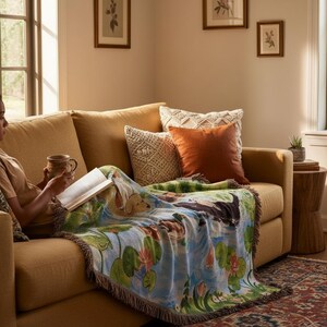 May include: A woven throw blanket featuring a colourful nature scene with ducks, lily pads, and flowers, draped over a mustard yellow sofa. The blanket has a fringed edge and displays a detailed illustration of waterfowl swimming in a pond.