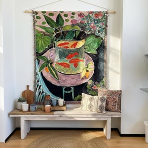 May include: A woven wall tapestry featuring a colorful painting of a round fishbowl filled with orange goldfish, resting on a pink table surrounded by green leaves and flowers. The tapestry has a fringed edge and is hung from a wooden rod.