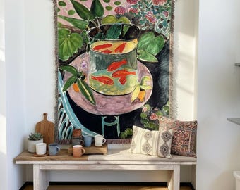 May include: A woven wall tapestry featuring a colourful painting of a round fishbowl filled with orange goldfish, resting on a pink table surrounded by green leaves and flowers. The tapestry has a fringed edge and is hung from a wooden rod.