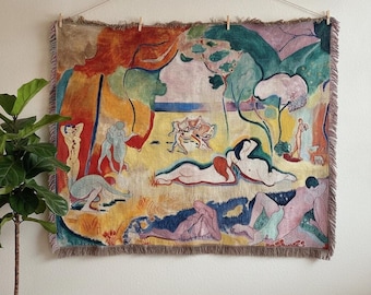 May include: A rectangular woven tapestry featuring a colourful landscape scene with multiple figures in various poses. The artwork displays shades of yellow, green, red, and blue, with a fringed border along the top and bottom edges, suspended by a string.