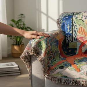 May include: A woven throw blanket featuring a colorful, abstract artistic pattern with prominent orange, blue, and green hues. The fabric has a textured, tapestry-like weave with frayed, fringed edges draped over the arm of a light gray chair.