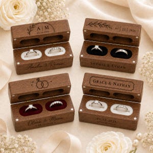 May include: Four rectangular dark wood ring boxes with hinged lids, each featuring custom laser-engraved names and dates. The boxes contain two circular slots lined with white or dark red velvet, holding silver-toned rings with clear stones.