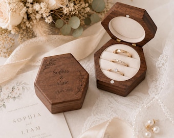 May include: Two hexagonal dark wood ring boxes with a hinged lid. One box is closed and engraved with the text Sophia & Liam and the date 07.09.2026. The open box contains three gold rings resting on a cream-colored velvet insert.