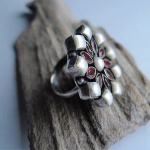 May include: A silver ring with a large, floral design. The ring features a cluster of white pearls, red gemstones, and square silver accents. The ring is set on a piece of driftwood, with a light blue background.
