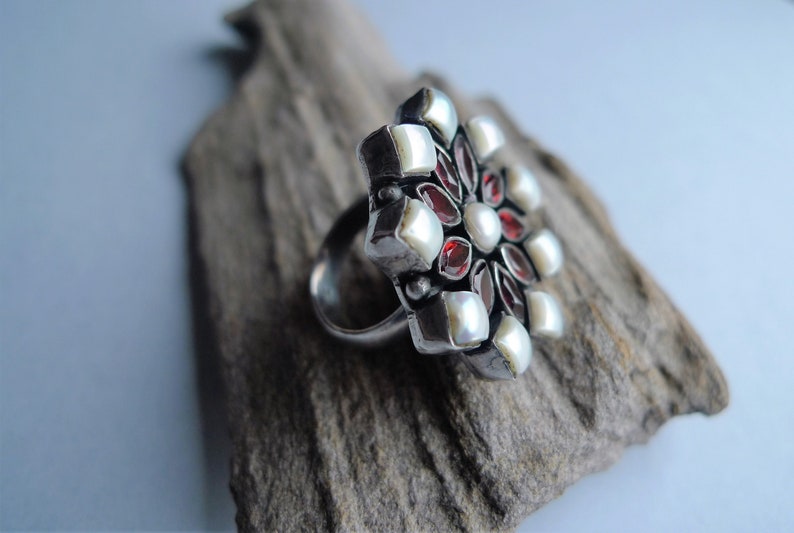 May include: A silver ring with a large, floral design. The ring features a cluster of white pearls, red gemstones, and square silver accents. The ring is set on a piece of driftwood, with a light blue background.
