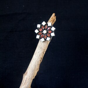May include: A flower-shaped ring with a silver setting. The ring features alternating red and white gemstones, with a pearl in the center. The ring is displayed on a piece of driftwood against a black background.