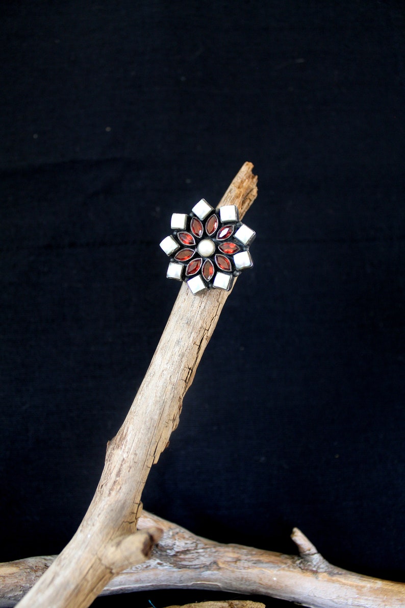 May include: A flower-shaped ring with a silver setting. The ring features alternating red and white gemstones, with a pearl in the center. The ring is displayed on a piece of driftwood against a black background.