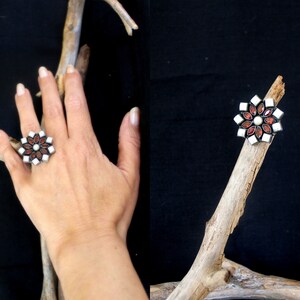 May include: A silver-toned ring with a floral design. The ring features a central white stone surrounded by red and white petal-shaped stones. The ring is displayed on a hand and a wooden branch against a black background.