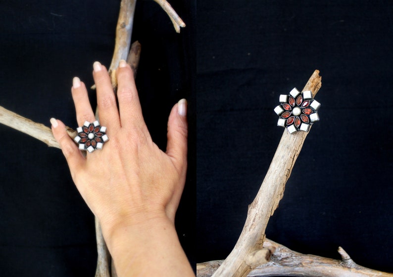 May include: A silver-toned ring with a floral design. The ring features a central white stone surrounded by red and white petal-shaped stones. The ring is displayed on a hand and a wooden branch against a black background.