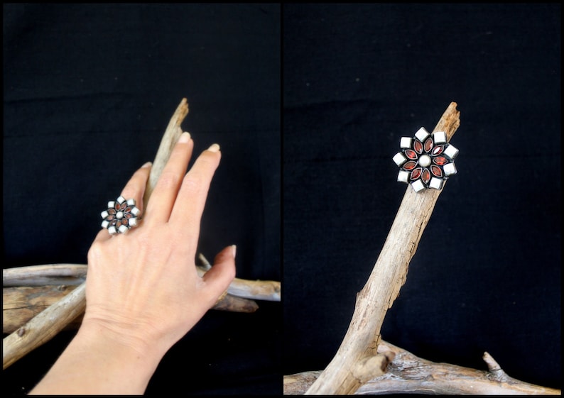 May include: A silver-toned ring with a floral design. The ring features alternating white and red gemstone petals surrounding a central pearl. The ring is displayed on a piece of driftwood against a black background.