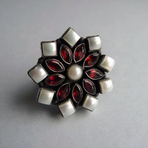 May include: A floral-shaped ring with a central pearl surrounded by red garnet petals and square white pearl accents. The ring has a dark metal setting and is set against a light gray background.