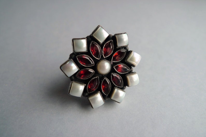 May include: A floral-shaped ring with a central pearl surrounded by red garnet petals and square white pearl accents. The ring has a dark metal setting and is set against a light gray background.