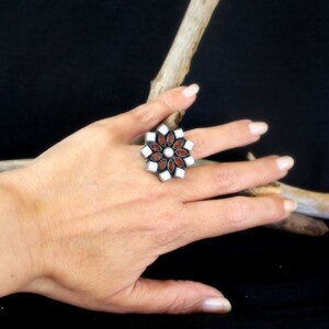 May include: A silver-toned ring with a floral design. The ring features alternating red and white stones, with a central white stone. The ring is displayed on a hand against a dark background.