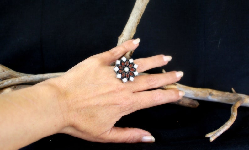 May include: A silver-toned ring with a floral design. The ring features alternating red and white stones, with a central white stone. The ring is displayed on a hand against a dark background.