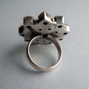 May include: Close-up of a silver ring with a large, ornate, flower-shaped setting. The setting has a textured surface with small, dark accents and a few light-colored stones. The ring band is smooth and rounded.