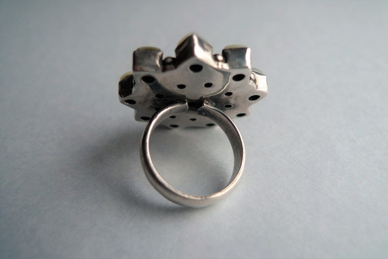 May include: Close-up of a silver ring with a large, ornate, flower-shaped setting. The setting has a textured surface with small, dark accents and a few light-colored stones. The ring band is smooth and rounded.