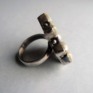 May include: A silver ring with a rectangular face adorned with several small, round, off-white pearls. The ring band is a simple, polished silver. The pearls are clustered together, creating a unique, eye-catching design.