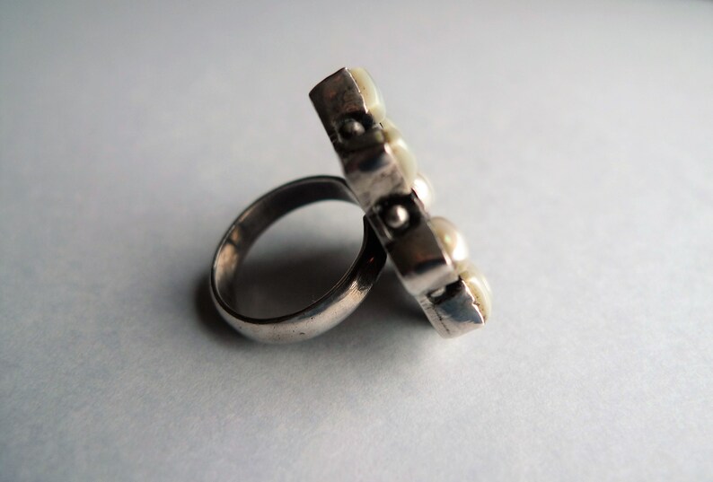 May include: A silver ring with a rectangular face adorned with several small, round, off-white pearls. The ring band is a simple, polished silver. The pearls are clustered together, creating a unique, eye-catching design.